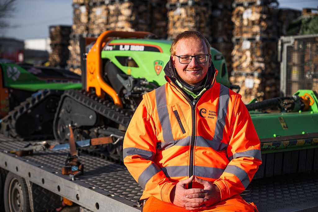 Lächelnder ELSTERWERK-Mitarbeiter in orange-gelber Warnschutzkleidung vor grüner Landschaftsbaumaschine - Portrait eines Garten- und Landschaftsbau-Fachmanns